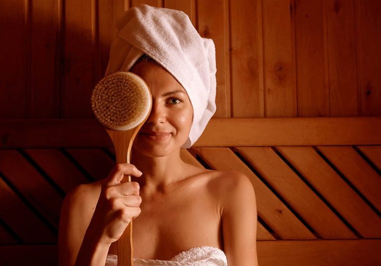 A young woman wearing a robe and relaxing in an infrared sauna while holding up a body brush to her face.