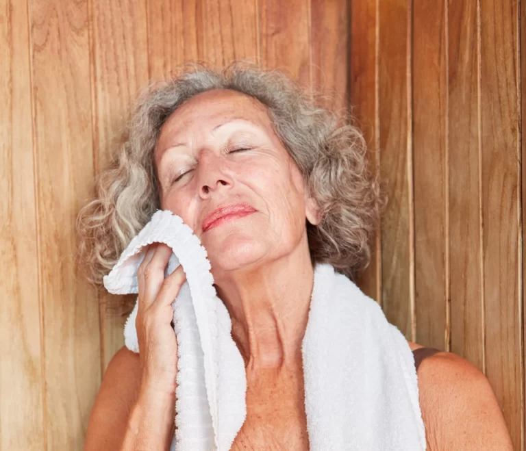 An older woman relaxing in an infrared sauna while blotting her face with a soft towel.